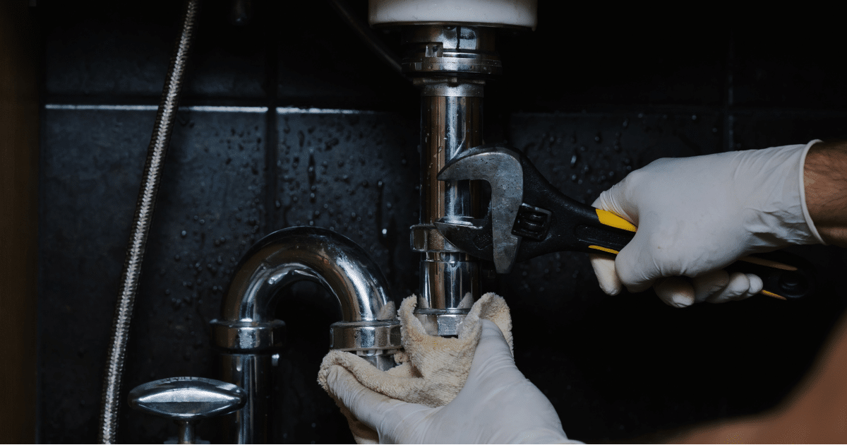 Plumber using an adjustable wrench to work on metal pipes beneath a bathroom sink.